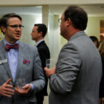 Two men in suits chat while holding drinks at a formal event. One wears a bow tie and name tag, while people in formal attire mingle in the background.