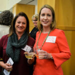 Two women smiling and holding drinks at an indoor event. They are both wearing name tags and standing in front of a wooden divider. Other individuals are visible in the background, creating a social and lively atmosphere.