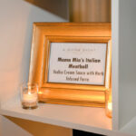A golden frame displays a sign for "Mama Mia’s Italian Meatball" with "Vodka Cream Sauce with Herb Infused Farro" written on it. The frame is positioned on a white shelf, flanked by two small lit candles.