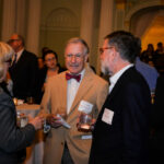 A group of people in formal attire are engaged in conversation at a social event. The setting is indoors, with warm lighting and decorative columns. Two men and a woman are visible in the foreground, holding drinks and wearing name tags.