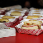 Open-faced sandwiches with ham and pickles are arranged in checkered paper trays on a red table. Napkins are stacked nearby.