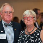 An elderly couple stands together at an event. The man wears a checkered shirt, bowtie, and glasses, with a name tag. The woman is in a black and white patterned dress, wearing red glasses. People are visible in the blurred background.