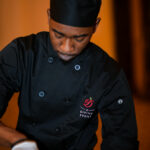 A chef in a black uniform and toque hat is focused on preparing food. He wears a black apron with a logo that reads "Divine Event." The background is softly blurred, highlighting the chef’s concentration and skill.