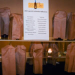 A table is set with several wine bottles wrapped in brown paper bags. A sign above reads, "AVLF is grateful to the Wine Wall donors," with a list of names below it. A candle is lit in the background.