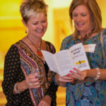 Two women stand together, smiling and reading a brochure titled "Event Map & Wine List." One woman holds a drink, and both are casually dressed, enjoying the event in a warmly lit setting.