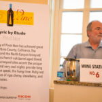 A man with a beard stands beside a "Wine Station No. 6" sign. Next to him, there's a display featuring wine tasting details for "Lyric by Etude Pinot Noir," along with a wine bottle graphic celebrating the 25th annual event.