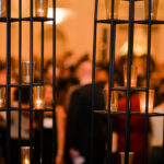 A decorative metal structure with lit candles in holders stands in the foreground, while a large gathering of people at a dimly lit event is blurred in the background. Warm colors dominate the scene, creating an inviting ambiance.