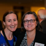 Two people smiling at an indoor event, one with short hair and glasses wearing a blazer with a name tag, and the other with long hair wearing earrings and holding a drink. Blurred background shows other attendees.
