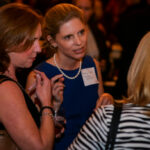 Three women are engaged in conversation at a social event. One holds a drink, and another wears a name tag. The background is blurred with soft lighting, suggesting a lively gathering.