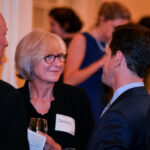 A group of people engaged in conversation at a social event. A woman with glasses and short blonde hair smiles while holding a glass. Two men, one with a bald head and the other with short dark hair, converse with her. People are blurred in the background.