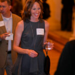 A woman in a gray dress smiling and holding a glass, standing near a display. A man in a tan suit is in the background. Both are wearing name tags. The setting appears to be an indoor social event.