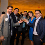 A group of five people, dressed in business attire, are smiling and holding drinks in a toast gesture at a social event. There are other attendees and elegantly decorated walls in the background.