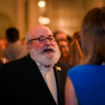 A bearded man with glasses is talking to a woman at a social gathering. The background is softly blurred, showing other people mingling in a warmly lit room.