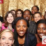 A group of smiling people posing in front of a shimmering gold sequin backdrop. They appear to be enjoying a celebratory event. The logos of "Smilebooth" and "avlf" are visible in the corners of the image.