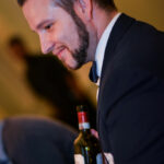 A man in a suit and bow tie holds a wine bottle. He's standing indoors, and the background is softly blurred, suggesting a social event or gathering.