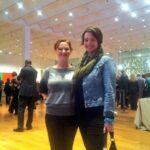 Two people standing and smiling in a large, well-lit room with wooden floors and modern decor. The scene includes other people mingling in the background, and one person is holding a purse.