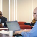 Two men are engaged in a conversation in an office setting. The man on the left, wearing a suit, is speaking, while the man on the right, in a blue shirt, types on a laptop. A paper cup and more people are visible in the background.