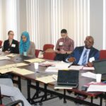 A group of people sit around a conference table covered with papers and laptops in a bright meeting room. One man is speaking while others listen attentively. Some participants have name badges, and one person is using a smartphone.