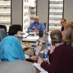 A group of people sit around a conference table in an office setting. Several individuals have notebooks and drinks. A person in a blue shirt is speaking, while others listen attentively. Large windows in the background reveal a cityscape.