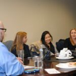 A group of people sitting around a conference table engaged in discussion. There are notebooks, papers, and plates with food on the table. The room has a neutral-colored wall.