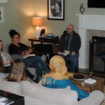 A group of people are seated around a coffee table in a cozy living room. Some are on a couch, while others sit in chairs. A fireplace is burning, and framed art hangs on the walls. Drinks and snacks are on the table, and they appear engaged in conversation.