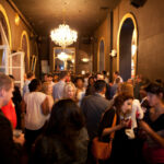 A crowded indoor gathering with people socializing in a dimly lit room. There are chandeliers, arched mirrors, and ornate decor. Some individuals hold drinks, creating a lively and engaging atmosphere.