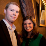 A man in a suit and a woman in a green top stand close together, smiling at the camera. The background has draped curtains and a mirror, suggesting an indoor event or social gathering.