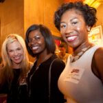 Three women are smiling and posing together in an indoor setting. They are standing close to each other, with warm lighting creating a friendly atmosphere. In the background, blurred figures and wall decor can be seen.