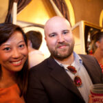 A smiling man and woman stand closely together at a social event. The man has a beard, and the woman has long hair. Both are casually dressed. People and party decorations are visible in the background.