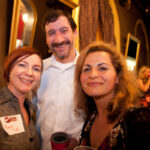 Three people smiling at a social gathering indoors. The two women are on either side of a man holding a drink. One woman has short red hair and a name tag. There are others conversing in the dimly lit background.