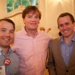 Three men are standing and smiling indoors at an event. They are wearing collared shirts and blazers. One man has a name tag with a button that says "Ask Me!" The background shows arched windows with soft lighting.