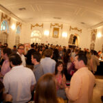A large group of people socialize at a formal indoor event. The room features elegant décor with tall windows and ornate moldings. Attendees are dressed in business casual attire, and some are holding drinks.