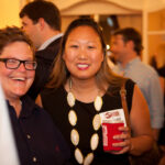Two people are smiling at a social event. One person holds a red cup and wears a large necklace. Others are gathered in the dimly lit background.