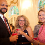Three smiling people holding red cups stand together indoors at an event. They are dressed in semi-formal attire. The setting features elegant decor with large windows and archways in the background.