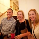 Three people smile while holding drinks indoors. The man on the left wears a checkered shirt, while the two women on the right wear light-colored tops. They stand near a staircase and framed photos on the wall.