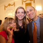 Three people pose together at an indoor event, smiling at the camera. The woman on the left is holding a green object, possibly food. The room has ornate decorations with arched windows and hanging drapes in the background.