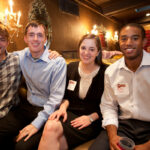Four people sitting on a couch in a warmly lit room with ornate decor. They are smiling, with two men on the left in shirts and two people on the right wearing name tags. The background features wall sconces and mirrors.