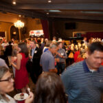A crowded indoor social event with people mingling, some holding drinks. Elegant chandeliers and dim lighting create a warm atmosphere. Attendees are dressed in business casual attire, and artwork is displayed on easels in the background.
