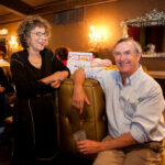 A man and woman are smiling and laughing in a dimly lit room with ornate decor, including mirrors and wall sconces. The man is seated on a couch and holding a drink, while the woman stands next to him with her arm on the couch.