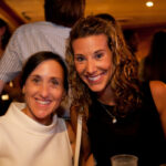 Two women smiling at the camera in a dimly lit setting. One has straight black hair and is wearing a white top, while the other has curly brown hair and is wearing a black top. People in the background appear blurred.