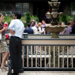 A woman and a man stand conversing by a decorative fountain during a social gathering. The scene is lively, with several people mingling in the background, dressed in formal attire. The area is adorned with greenery and small lights.