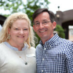 A smiling woman with curly blonde hair wearing a light-colored top stands next to a man with short dark hair and glasses in a checkered shirt. They are outdoors, with trees and a building in the background.
