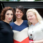 Three people smiling and standing closely together. The person on the left has curly hair, the person in the middle has short hair, and the person on the right has blonde hair and is holding a drink. They appear to be outdoors.