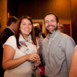 A woman and a man smiling at a gathering. The woman is holding a drink and wearing a white top with a scarf. The man is wearing a light-colored button-up shirt. They are in a warmly lit room with several other people in the background.