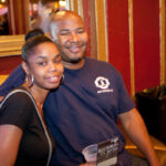 A man and a woman are sitting on a gold couch in a dimly lit venue. The man is wearing a blue t-shirt and smiling, holding a drink. The woman is in a black top, sitting close and relaxed. A mirror and red walls decorate the background.