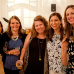 A group of five women smiling and standing closely together at an indoor gathering. They are holding drinks and dressed in casual, colorful clothing, with one wearing a name tag. The room has a bright, elegant setting with white paneled walls.