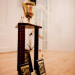 A tall trophy with gold accents and a figurine on top stands on a wooden floor. Three participant plaques are placed in front of it. The background features a white wall with an arched window.
