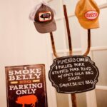 Two caps are displayed on a stand next to a sign listing menu items like pimento cheese and pulled pork. Another sign reads "Smoke Belly BBQ Parking Only," featuring a pig graphic. The setup has a rustic feel with a white wall in the background.