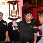 A man in a black shirt and cap holds up a trophy on stage, celebrating with a group of people. A large screen in the background displays text related to the event.