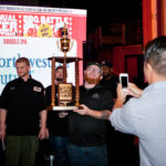 A group of people stand on stage at a BBQ event. One person in the center holds up a large trophy, smiling as someone takes a photo with a phone. A screen in the background displays "Annual BBQ Battle" and other text.